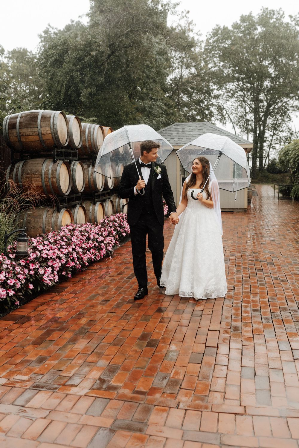 Bride and groom walk hand-in-hand under clear umbrellas on a rainy brick path beside wine barrels and pink flowers.