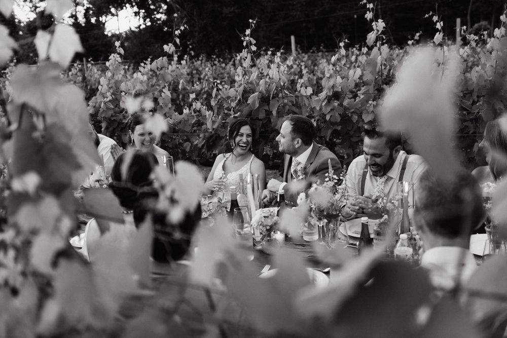 Black and white photo of guests laughing and dining together at an outdoor wedding reception surrounded by lush greenery.