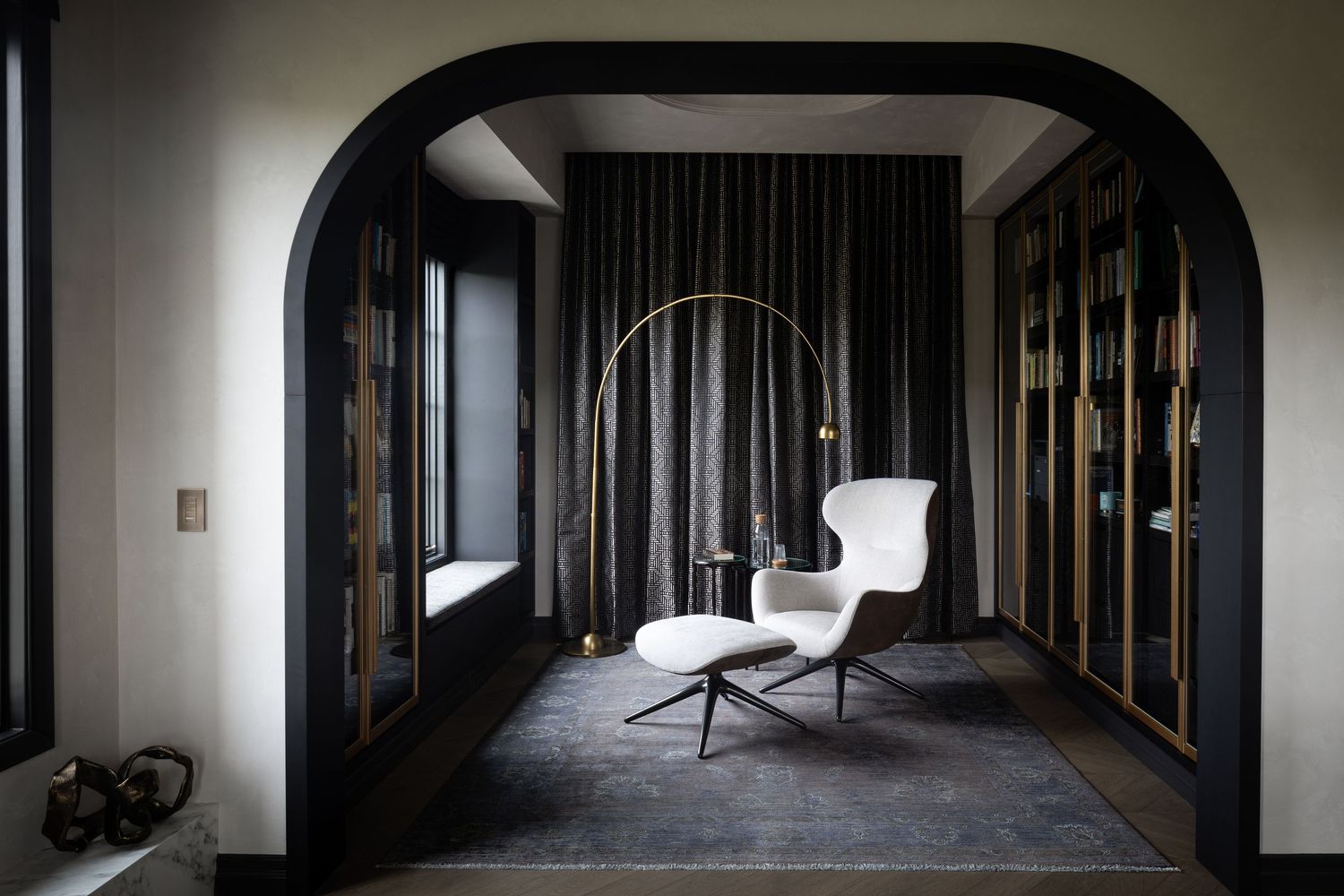 Elegant dark interior with arched doorway, white lounge chair, ottoman, arc floor lamp, and gold-framed glass cabinets.