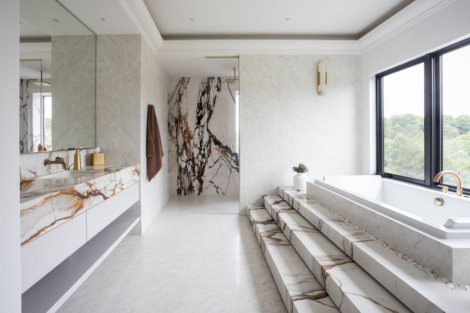 Luxurious white marble bathroom with gold fixtures, freestanding soaking tub, double vanity, and large window overlooking greenery.