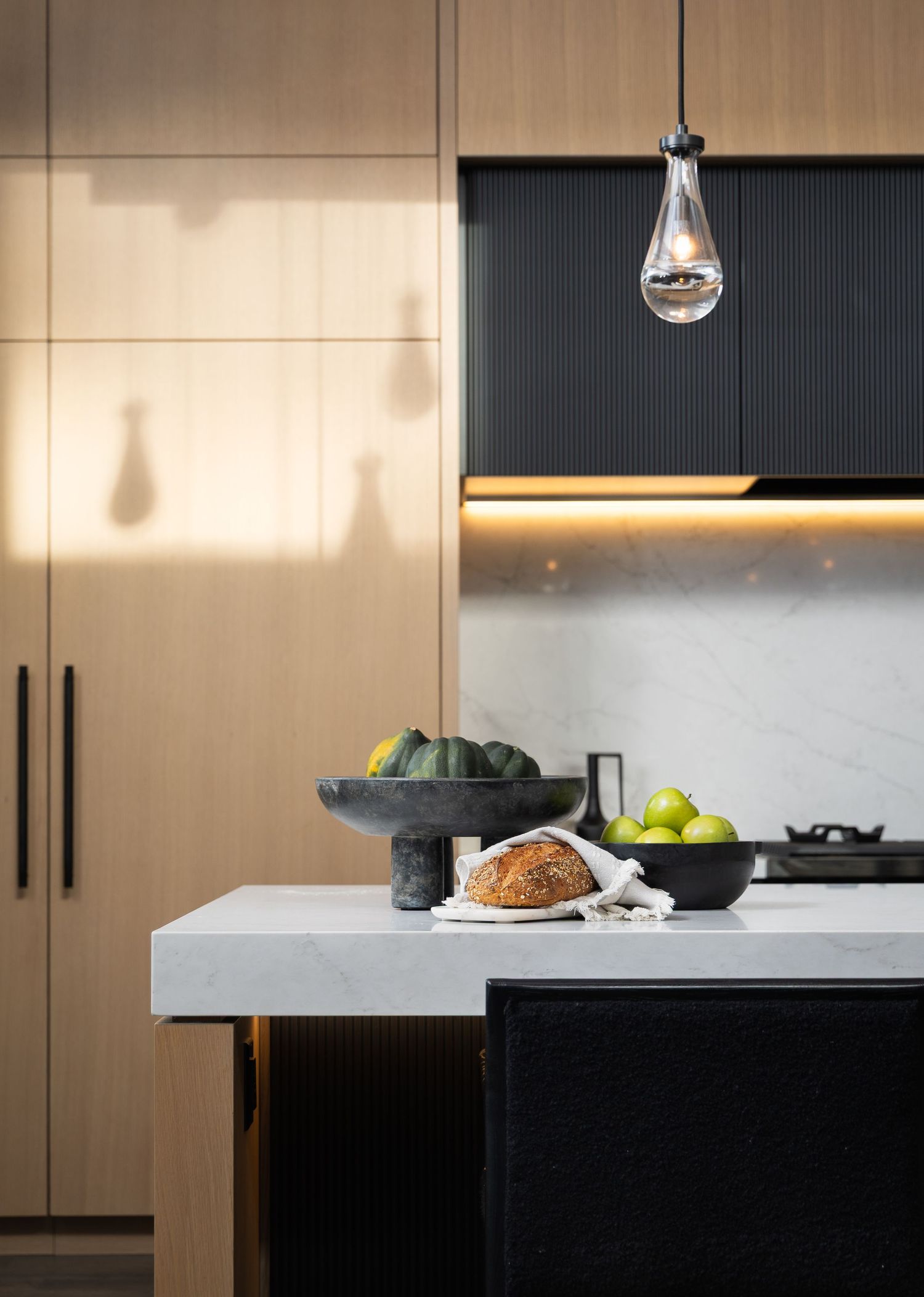 Modern kitchen island with marble countertop, black cabinetry, wood panels, pendant light, and fresh fruit display.