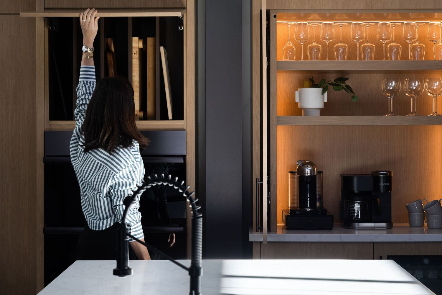 Woman reaching into dark kitchen cabinet beside illuminated open shelving with glassware and coffee machine.