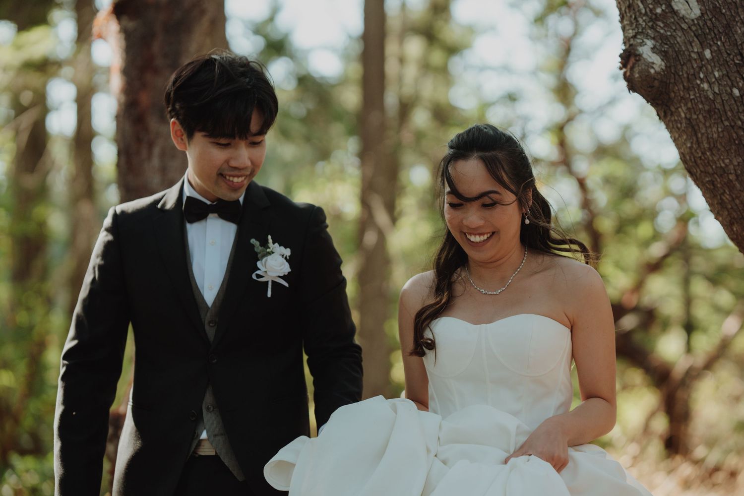 Smiling bride in white gown walks with groom in black tuxedo through sunlit forest during wedding photo session. Victoria, BC, Vancouver Isl