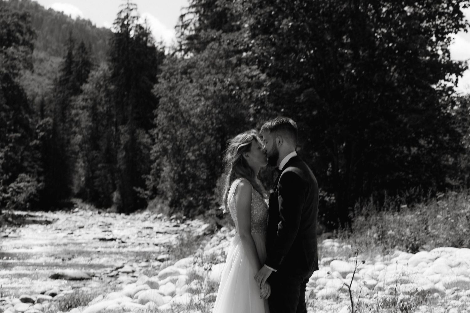 A black and white wedding photo of a couple by a riverbank surrounded by trees.