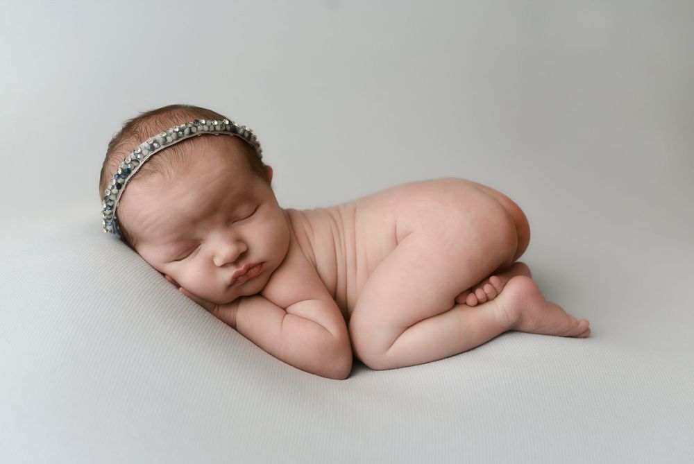 Peaceful newborn baby girl sleeping curled up, wearing a delicate rhinestone headband, on a soft white background.