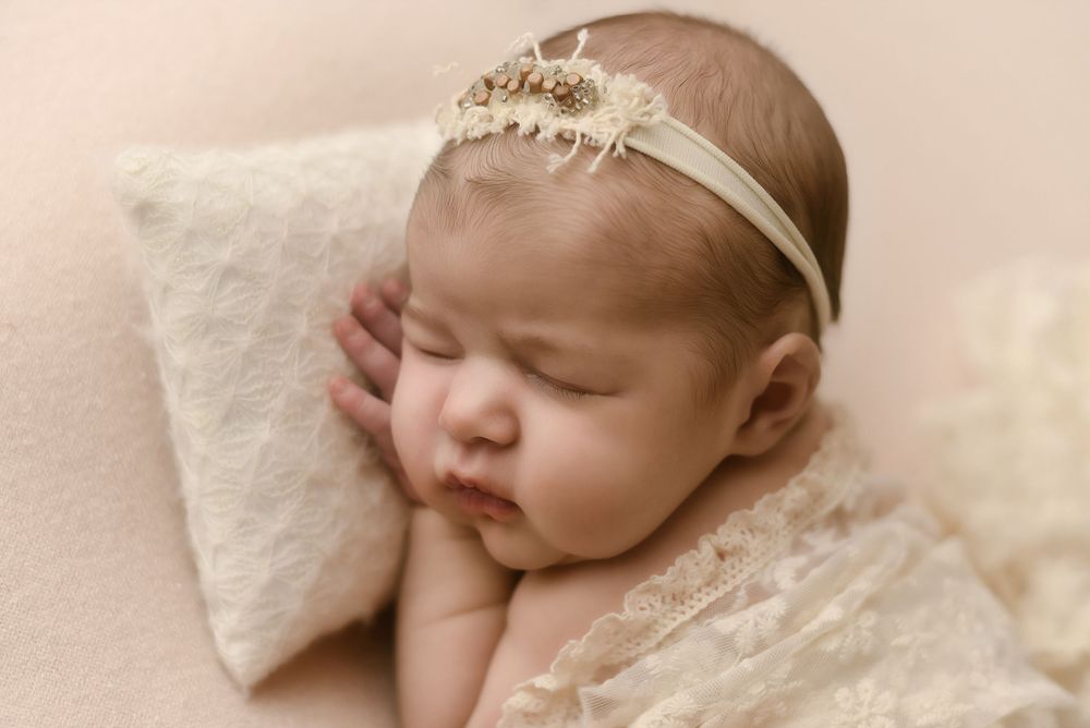 Peaceful newborn baby girl sleeping on cream lace wrap wearing delicate pearl headband in soft pink studio setting.
