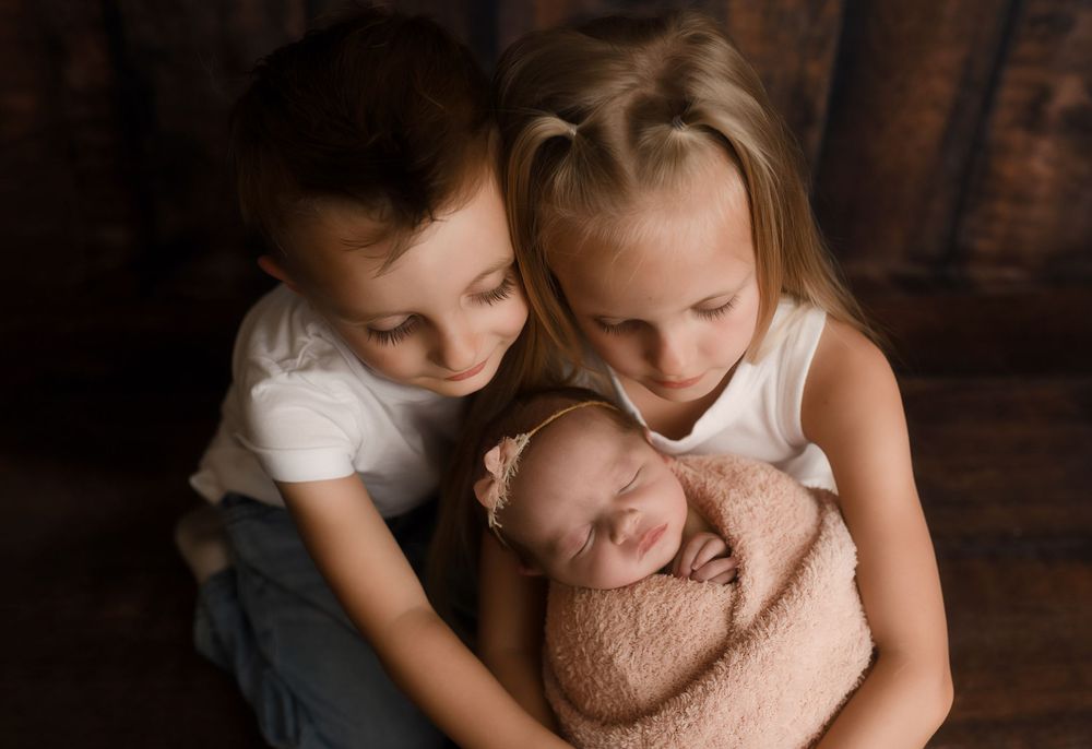 Two children tenderly hold a sleeping newborn baby wrapped in a soft pink blanket in a warm studio portrait.