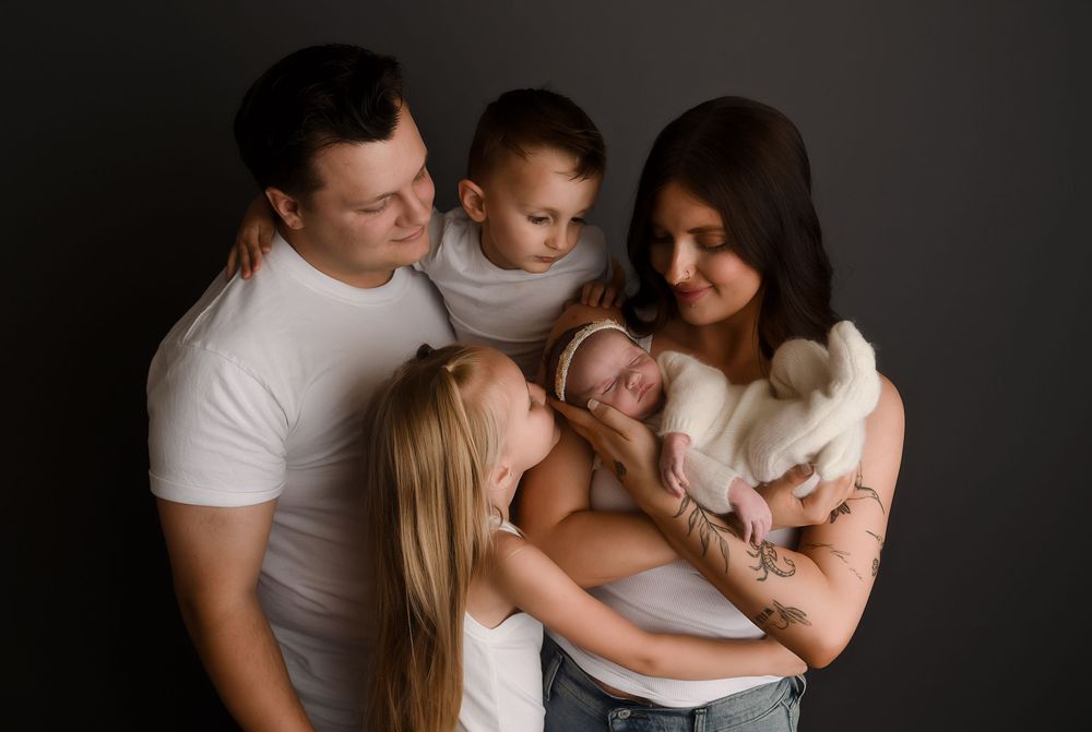 Family of five in white tops poses with newborn baby against dark background in professional studio portrait.