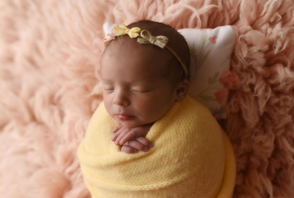Newborn baby swaddled in yellow wrap with gold floral headband sleeping on pink fluffy backdrop.