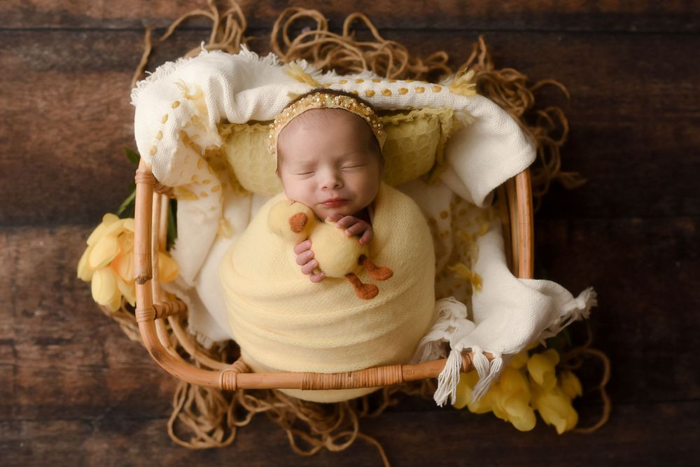 Newborn baby swaddled in yellow wrap sleeping in wicker basket with soft blanket and small duck prop.