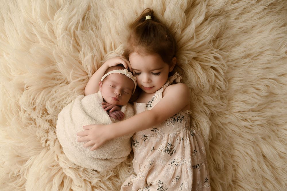 Young girl in floral dress tenderly holds swaddled newborn baby on fluffy cream fur rug.