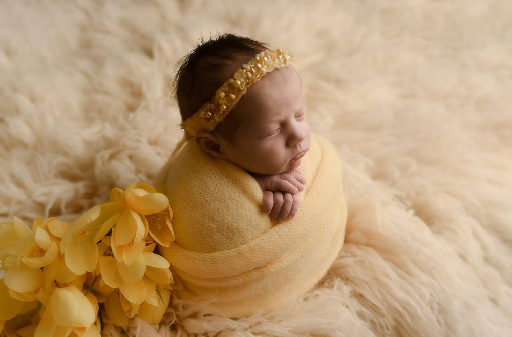 Sleeping newborn baby wrapped in yellow swaddle with gold headband on fluffy cream rug with yellow flowers.