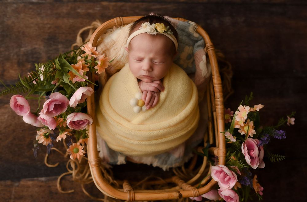 Newborn baby sleeping peacefully in a wicker basket wrapped in yellow, surrounded by pink flowers.