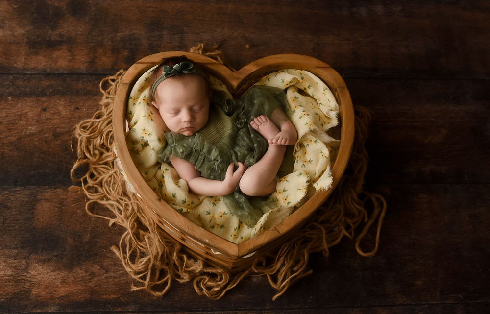 Newborn baby sleeping peacefully in a heart-shaped wooden bowl with green wrap and floral headband on rustic wood.