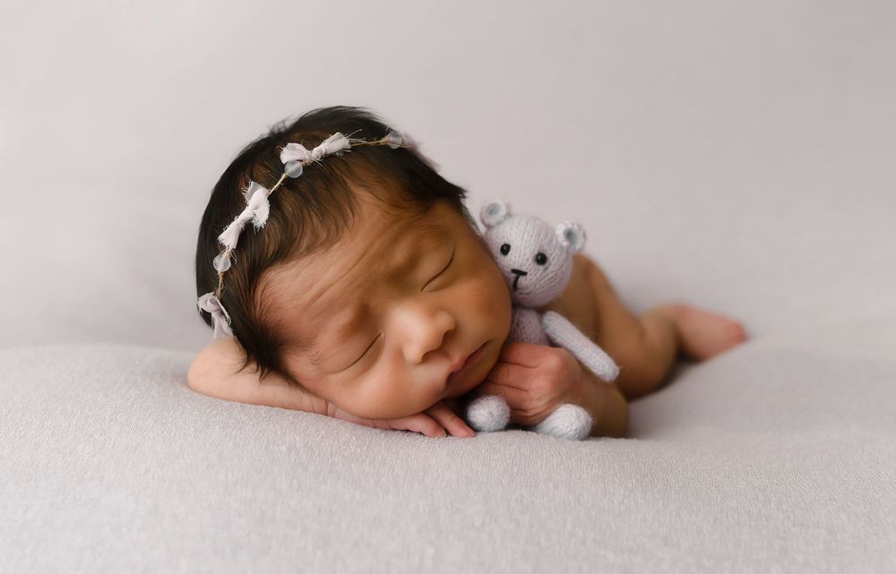 Sleeping newborn baby girl wearing a white bow headband, cuddling a tiny knit teddy bear on a soft gray blanket.