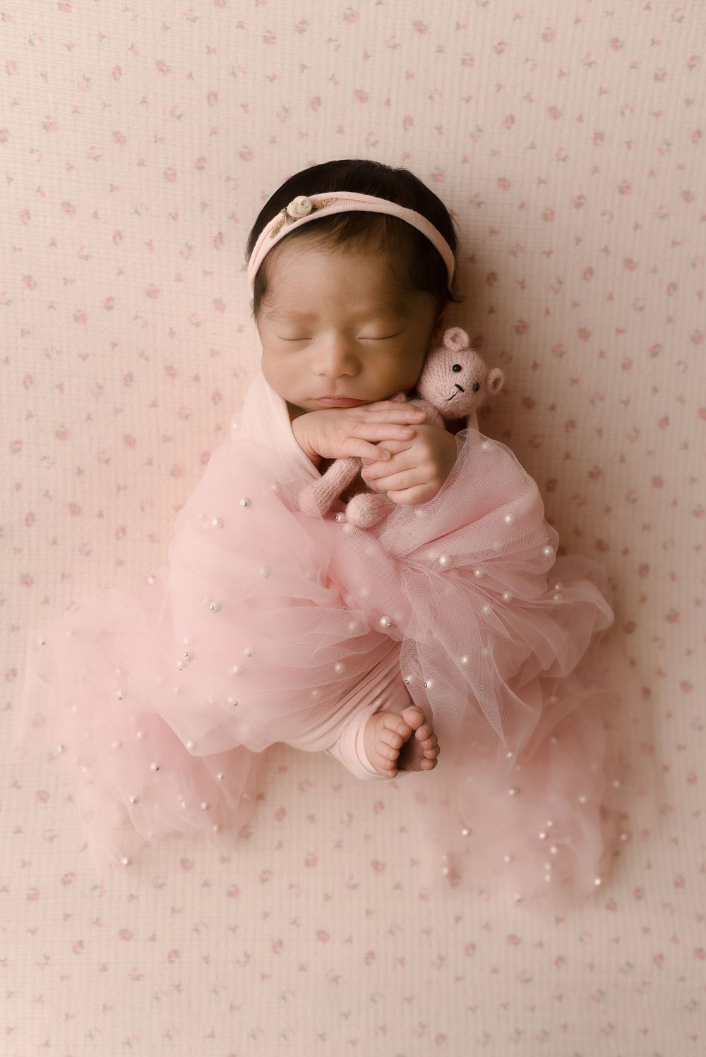 Newborn baby girl wrapped in pink pearl tulle, holding a tiny bear, wearing a pink headband, sleeping peacefully.