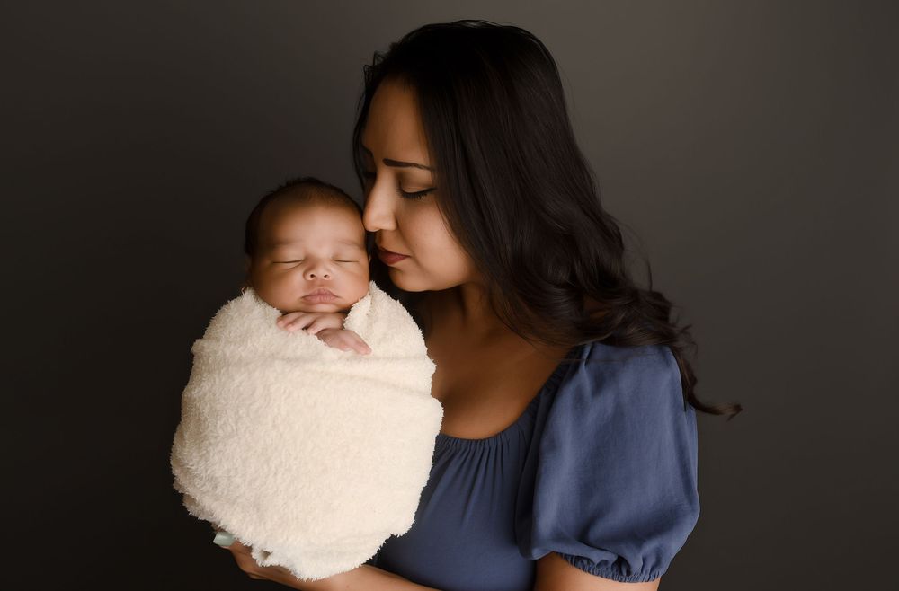 A mother tenderly kisses her sleeping newborn baby wrapped in a cream swaddle blanket.