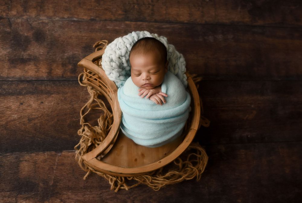 Sleeping newborn baby swaddled in a soft teal wrap, nestled in a wooden bowl on a rustic dark wood background.