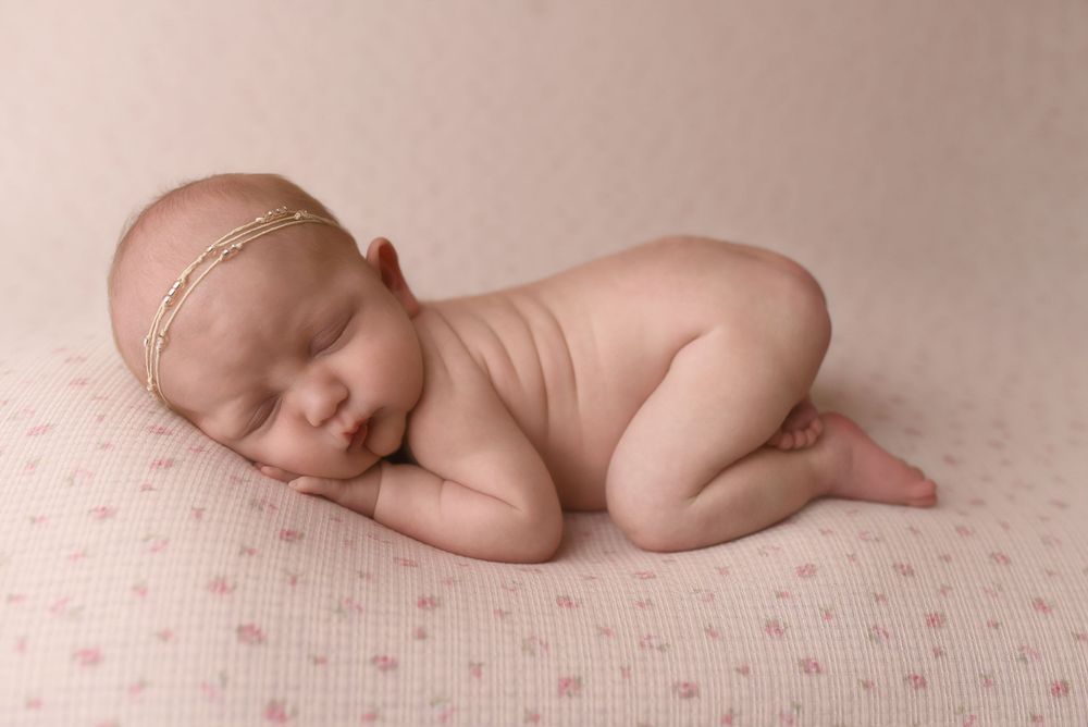 Newborn baby girl sleeping peacefully on a pink polka dot blanket, wearing a delicate gold headband.