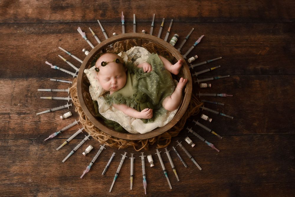 Newborn baby sleeping peacefully in a wooden bowl wrapped in green, surrounded by syringes in a sunburst pattern.