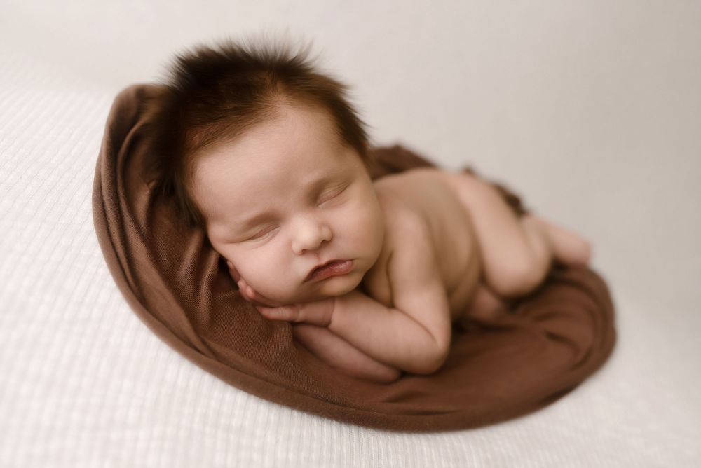 Peaceful newborn baby sleeping curled up on a brown wrap in a bowl pose during a professional newborn photography session.