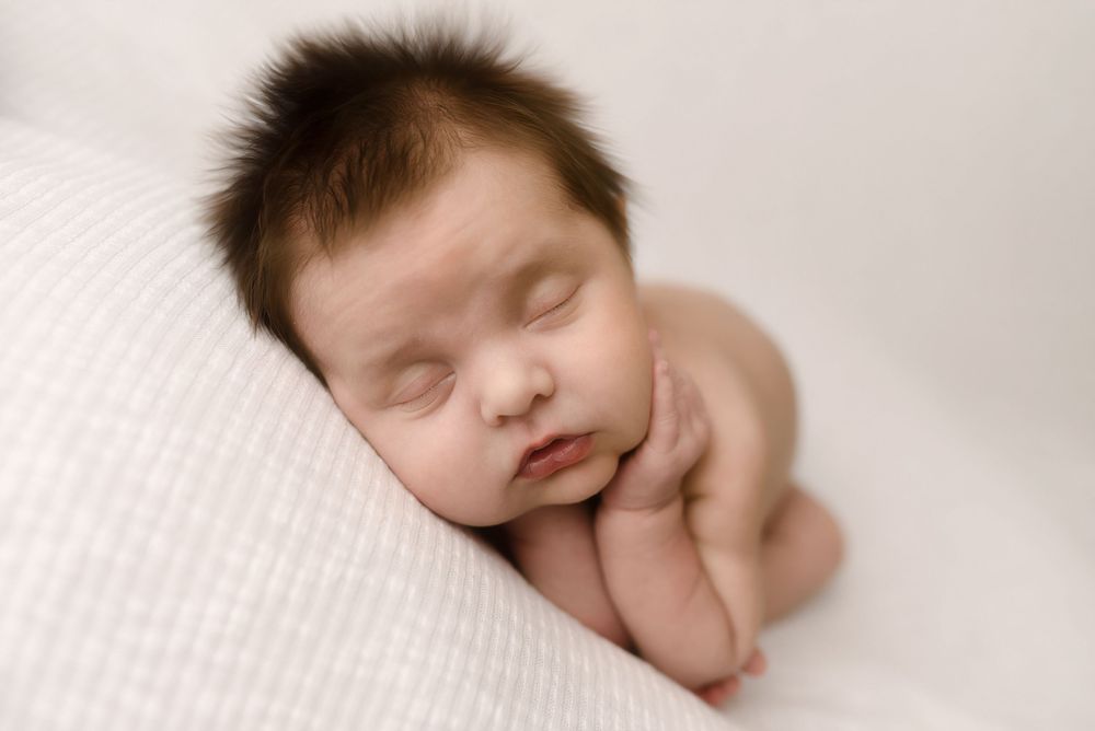 Peaceful newborn baby with dark hair sleeping curled up on a soft white blanket in a professional photo.