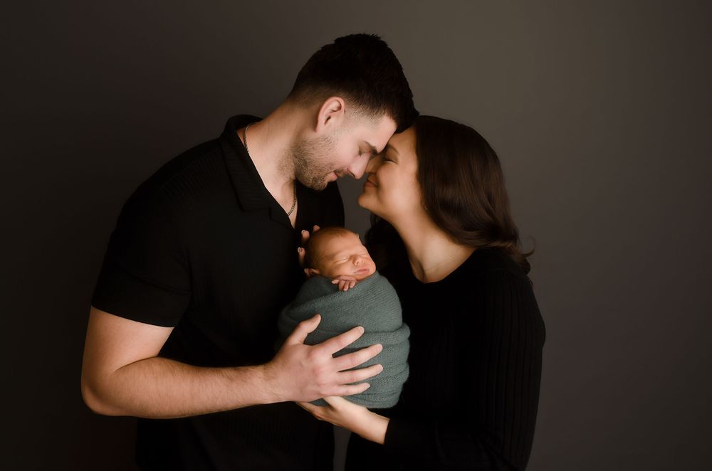 Parents tenderly kiss their swaddled newborn baby in a professional studio portrait photo.