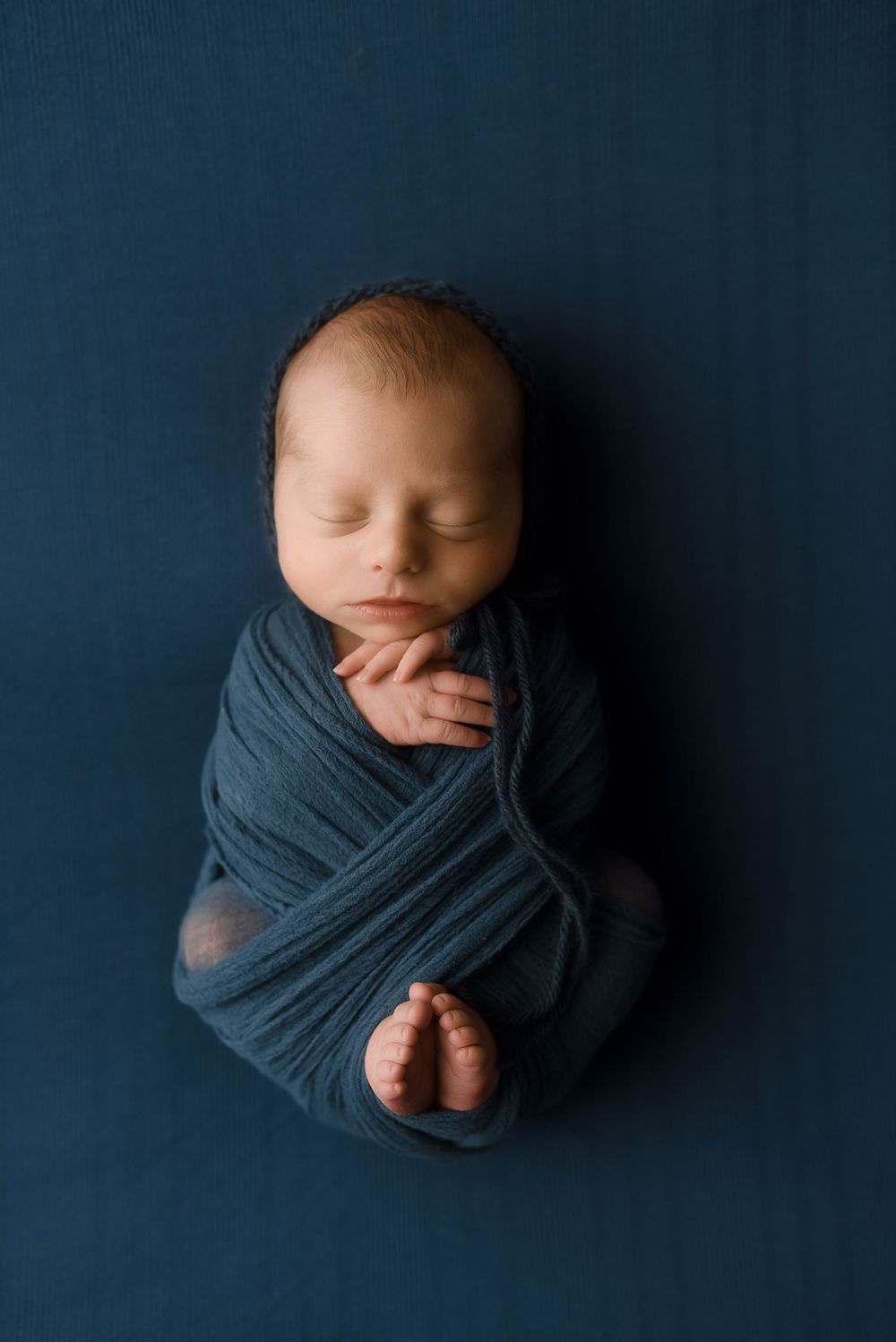 Newborn baby swaddled in a teal wrap, peacefully sleeping on a navy blue background.