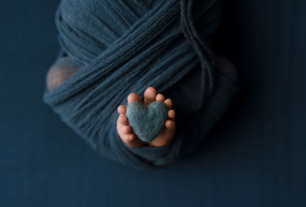 Newborn baby feet holding a teal felt heart, wrapped in soft blue yarn for a cozy newborn photo.