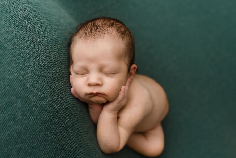 Peaceful newborn baby sleeping curled up on a green backdrop in a professional newborn photography session.