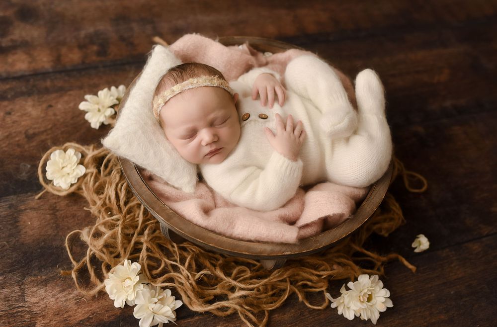 Sleeping newborn baby in white outfit and pink headband posed in wooden bowl with cream flowers.