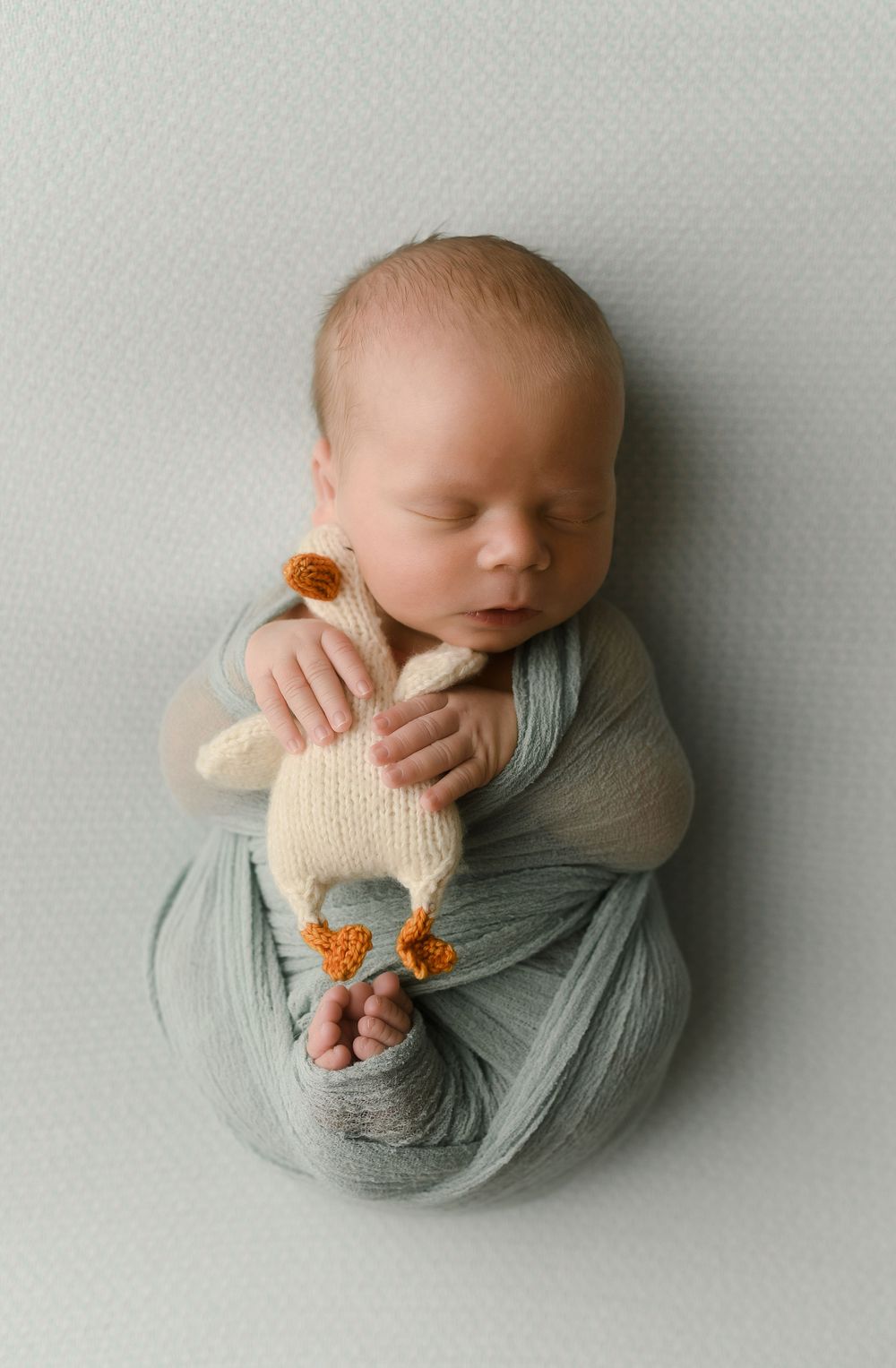 Sleeping newborn baby swaddled in soft teal wrap, gently holding a small knitted stuffed animal toy.