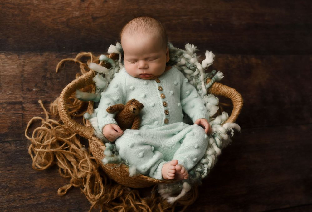 Newborn baby sleeping in a rustic basket wearing a mint green outfit, holding a tiny teddy bear on a wooden background.