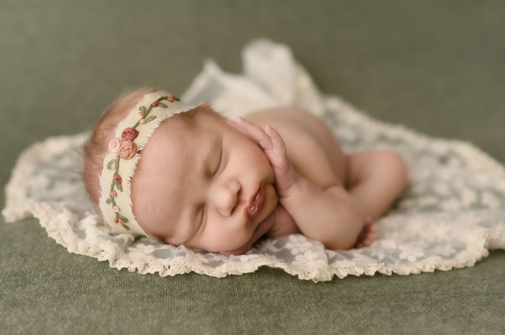 Newborn baby girl sleeping peacefully on lace blanket wearing floral headband on green background.