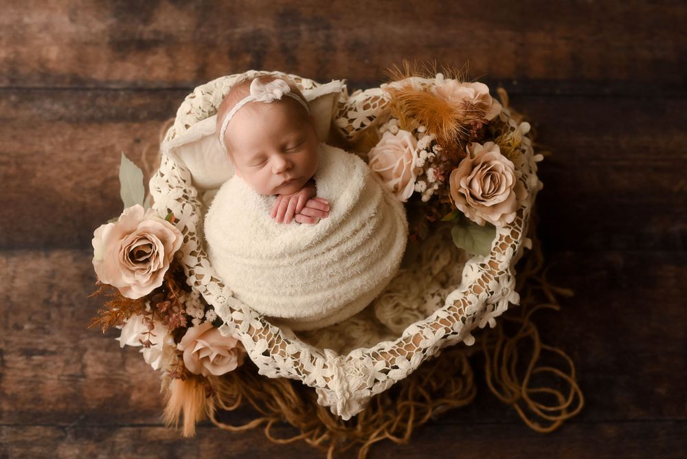 Newborn baby wrapped in cream knit, sleeping in heart-shaped basket with roses on wood backdrop.