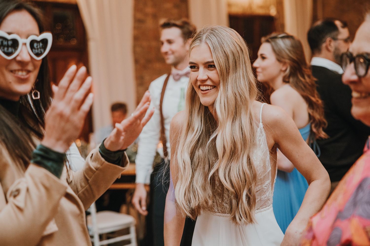 Bride with long blonde hair smiling and dancing with guests at a lively wedding reception celebration.