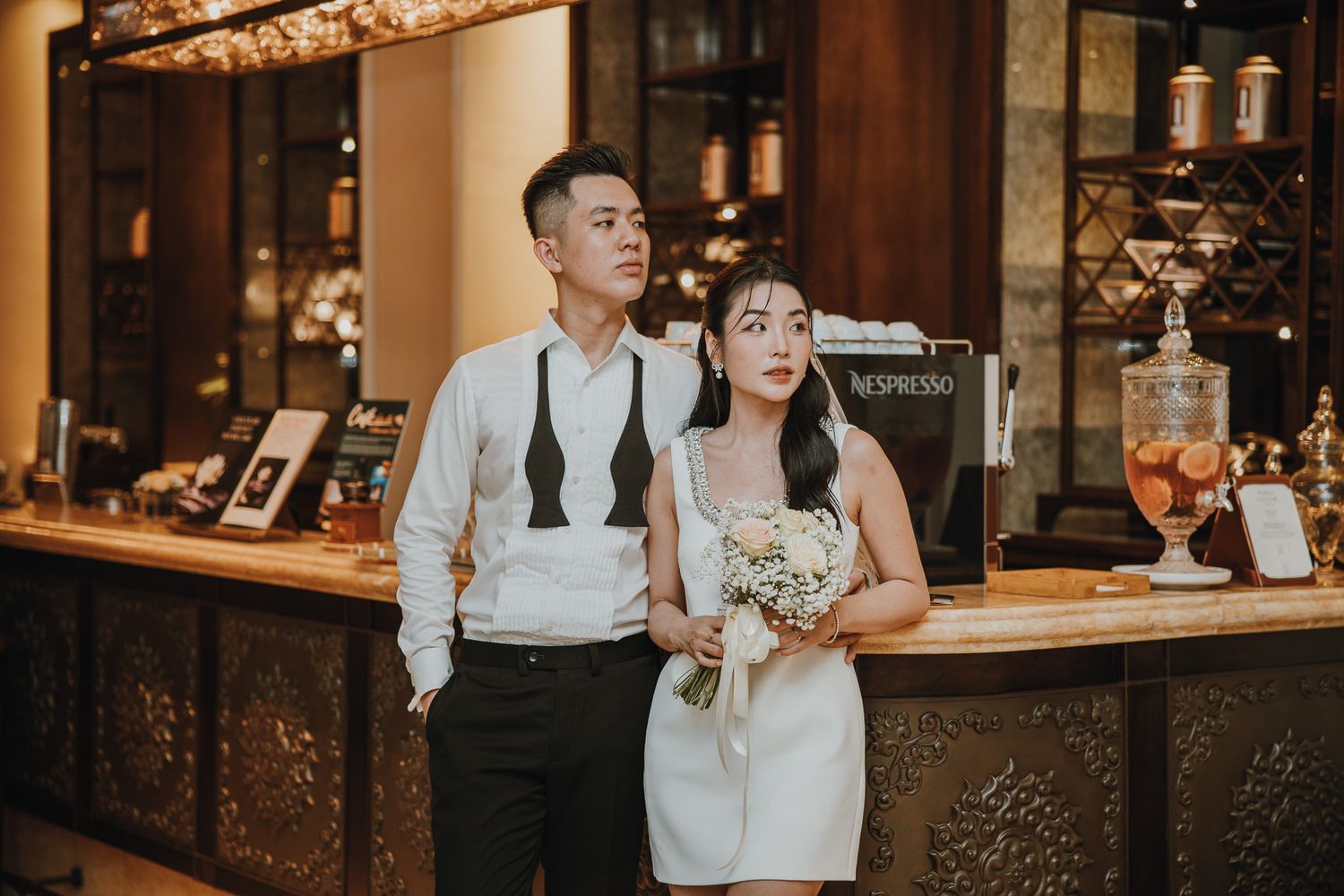Couple in elegant attire poses at a bar, woman holding a bouquet in a white dress, man in white shirt with suspenders.