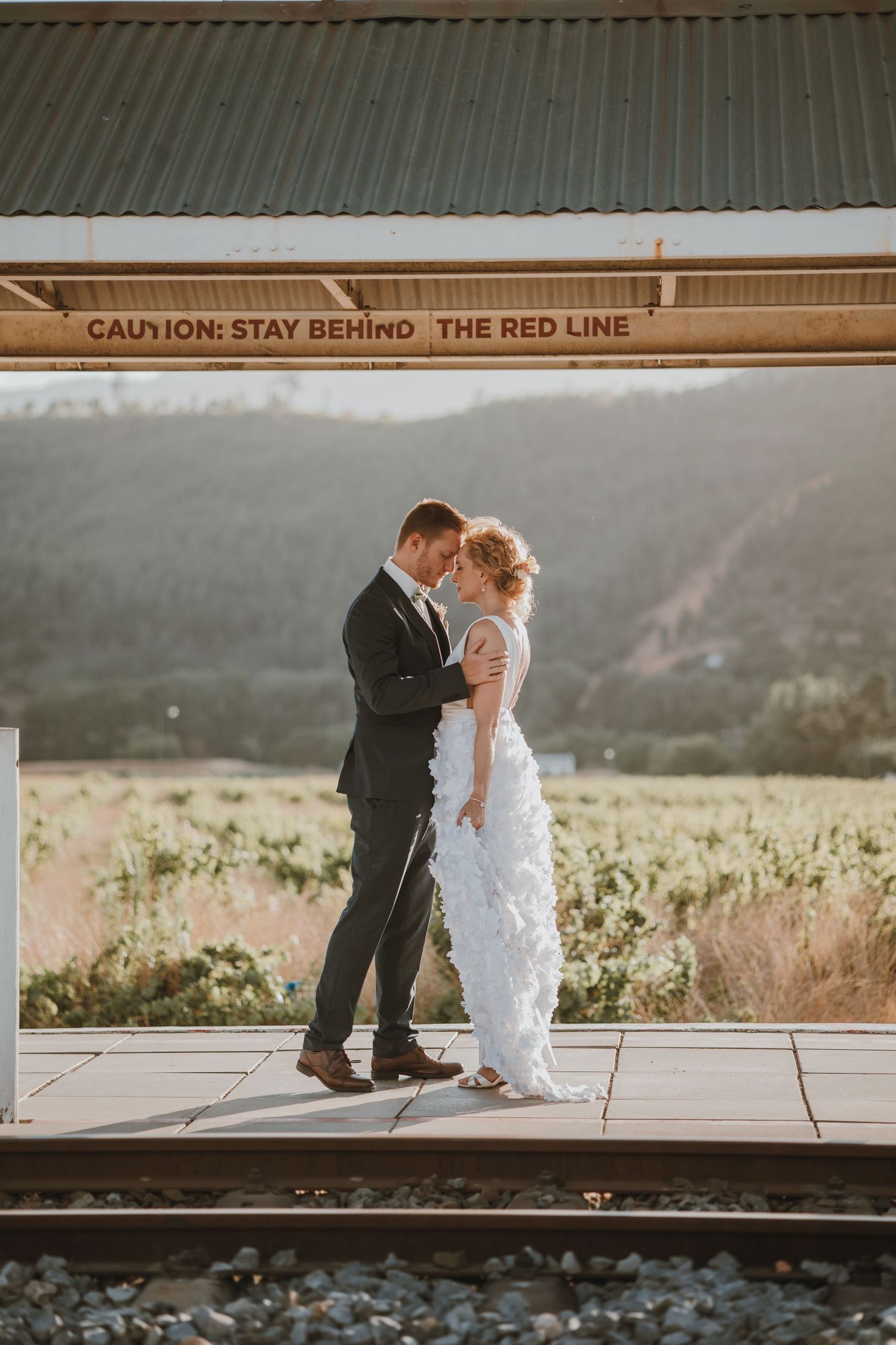 Bride and groom share a romantic moment on a train platform with a scenic vineyard backdrop at golden hour.