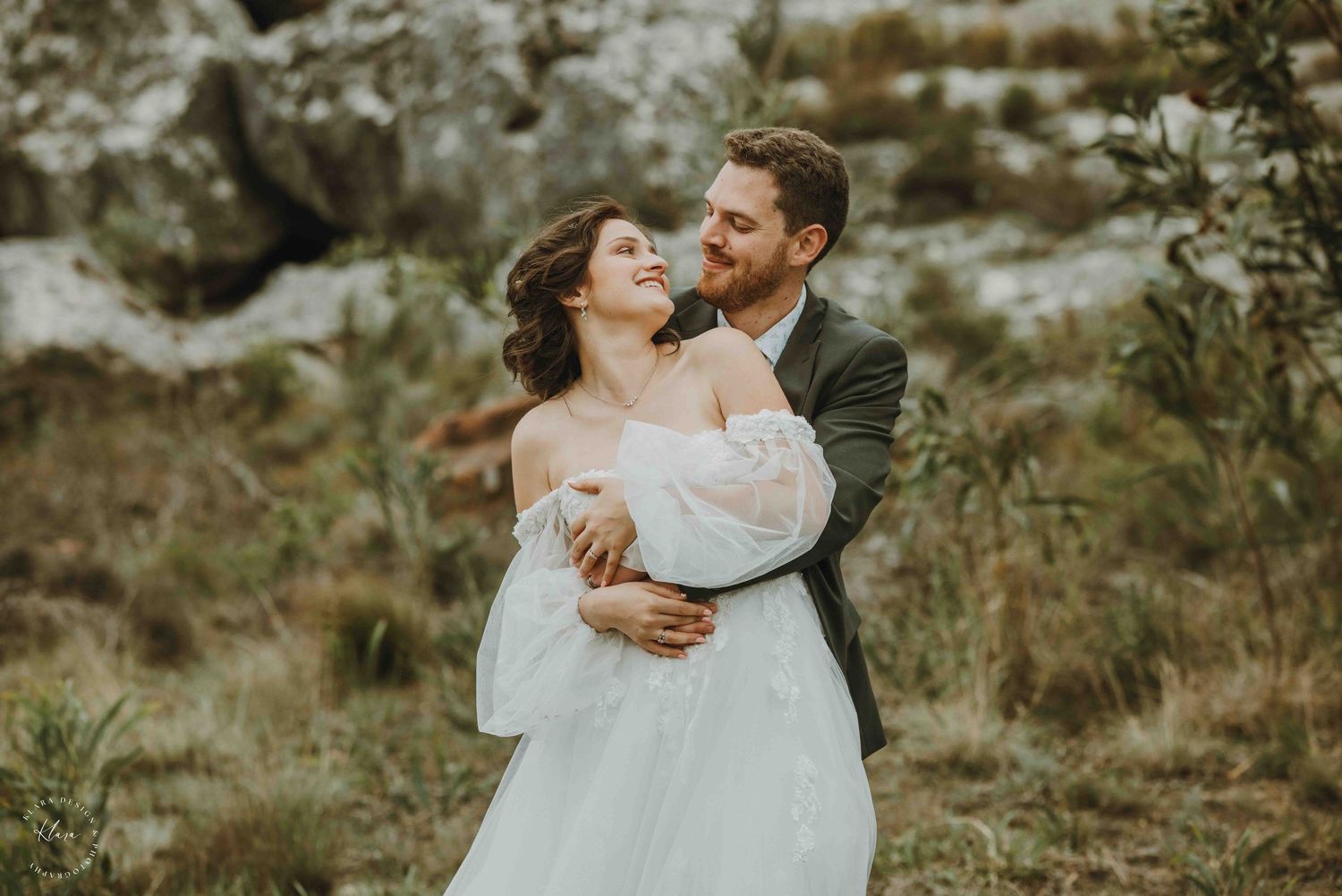 A couple embraces in a romantic outdoor wedding shoot, the bride in a white gown and groom in a gray suit among wild greenery.