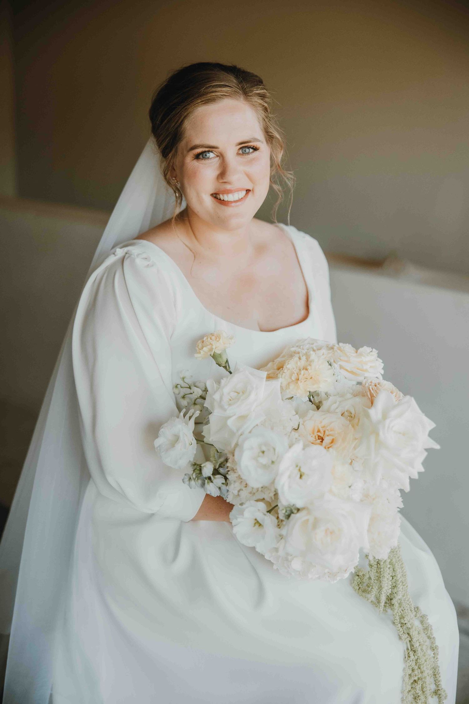Smiling bride in white long-sleeve gown holding lush ivory floral bouquet, wearing veil, seated indoors.