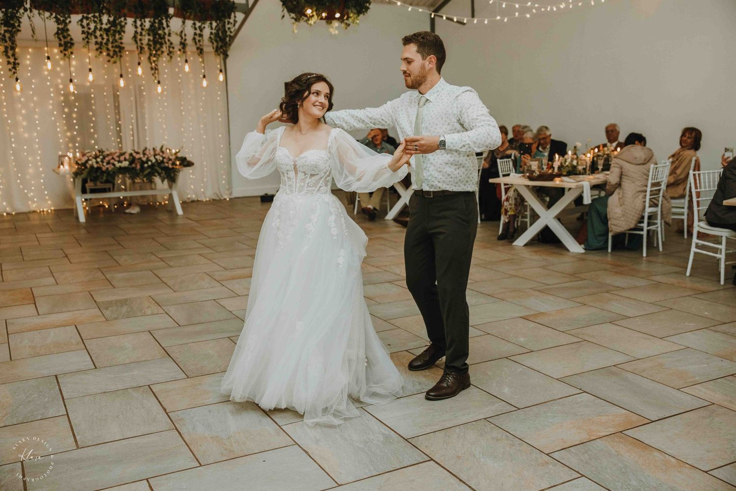 Bride and groom share their first dance in a decorated wedding venue with string lights and floral arrangements.
