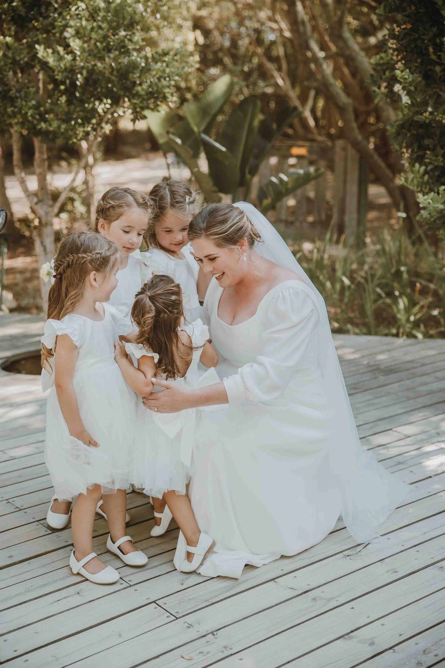 Bride in white gown kneeling on wooden deck with four flower girls in white dresses, sharing a tender moment outdoors.