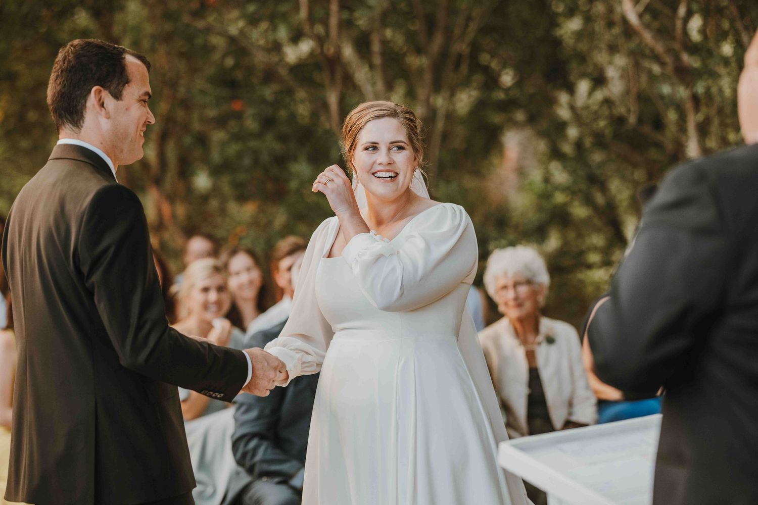 Bride in white gown laughing joyfully while holding groom's hand during outdoor wedding ceremony with guests seated behind.