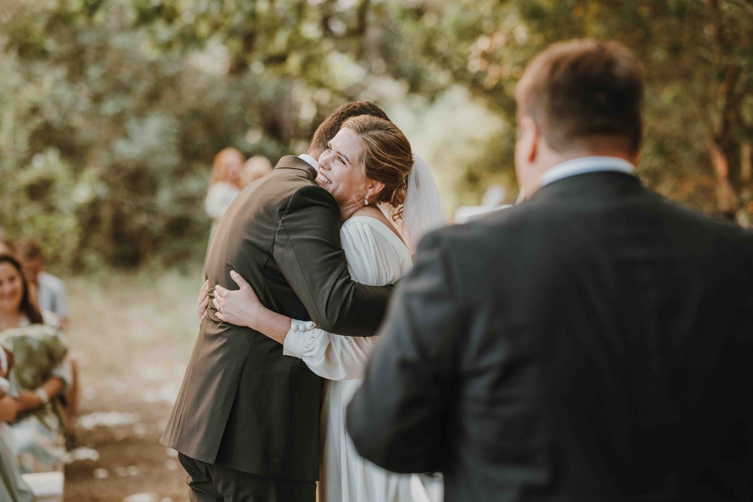 Emotional wedding ceremony hug between bride and guest outdoors, with groom in gray suit watching in foreground.