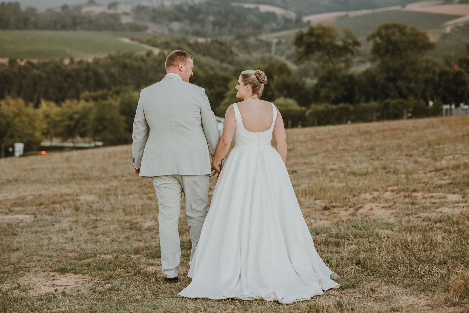 A bride and groom walk hand in hand through a scenic green hillside field on their wedding day.