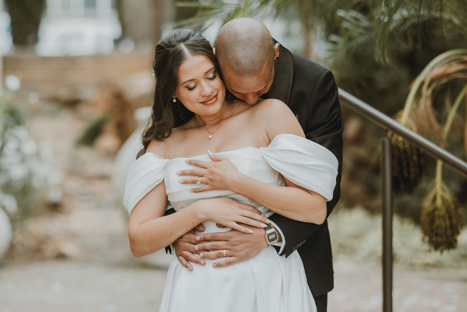Bride in off-shoulder white gown embraced from behind by groom in black suit during outdoor wedding portrait.