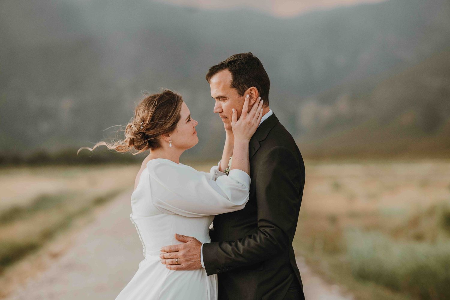Bride in white dress tenderly holds groom's face outdoors with misty mountains in background.