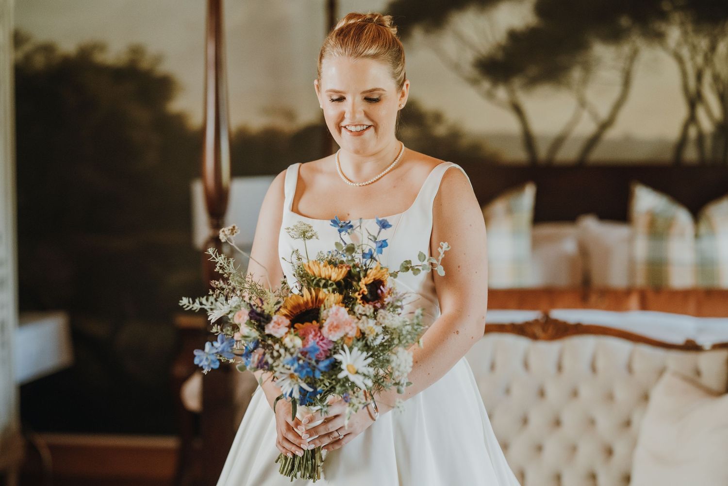 Bride in white gown smiling at colorful wildflower bouquet with sunflowers and daisies indoors.