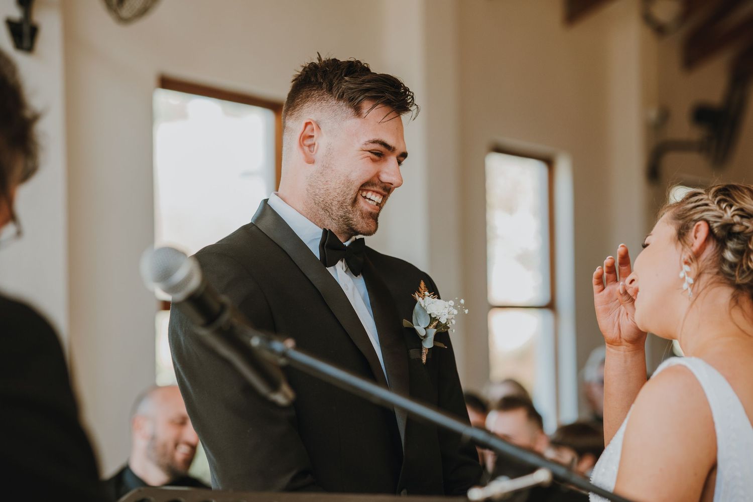 Groom in black tuxedo smiling joyfully at bride during wedding ceremony at elegant venue.