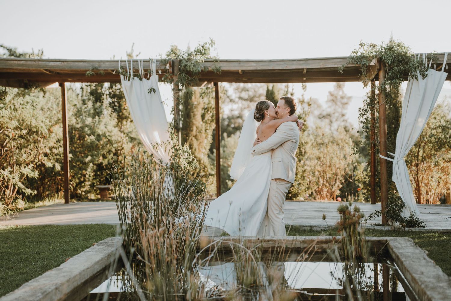 Bride and groom share first kiss under rustic wooden arch with white drapes and greenery at outdoor wedding ceremony.